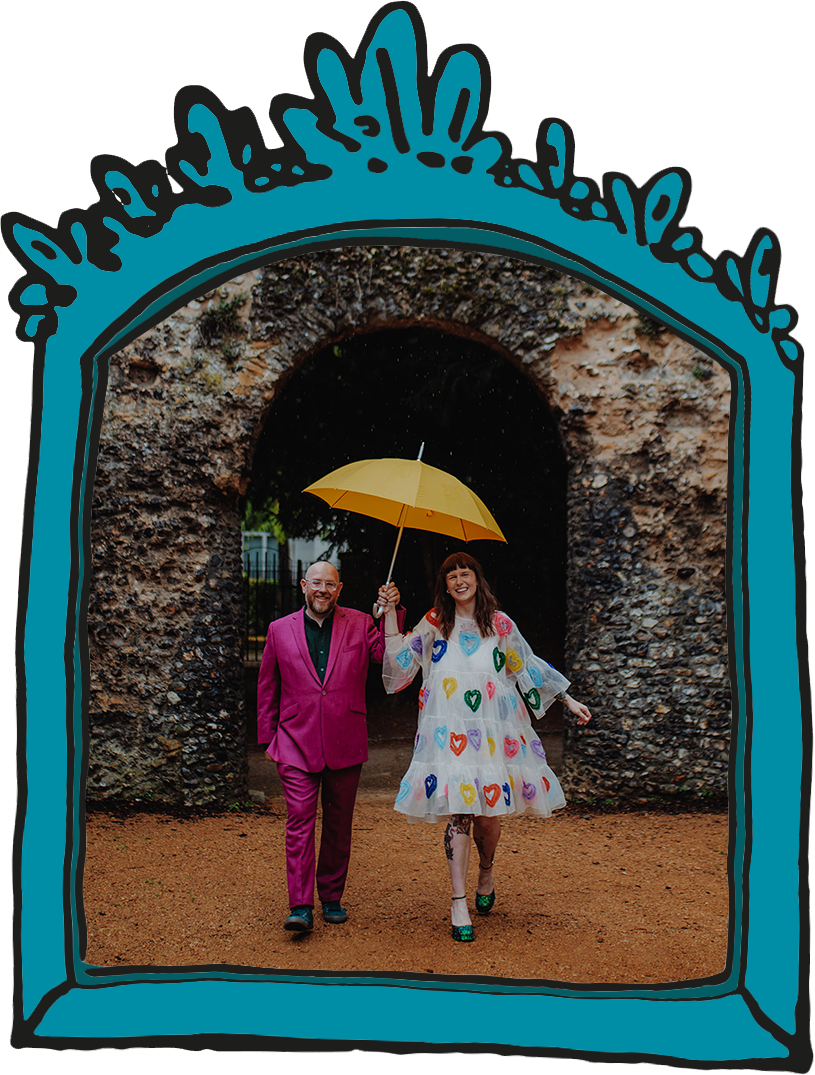 Groom in bright pink suit and bride in knee length dress with rainbow hearts on. Stood under bright yellow umbrella, in front of ruins.