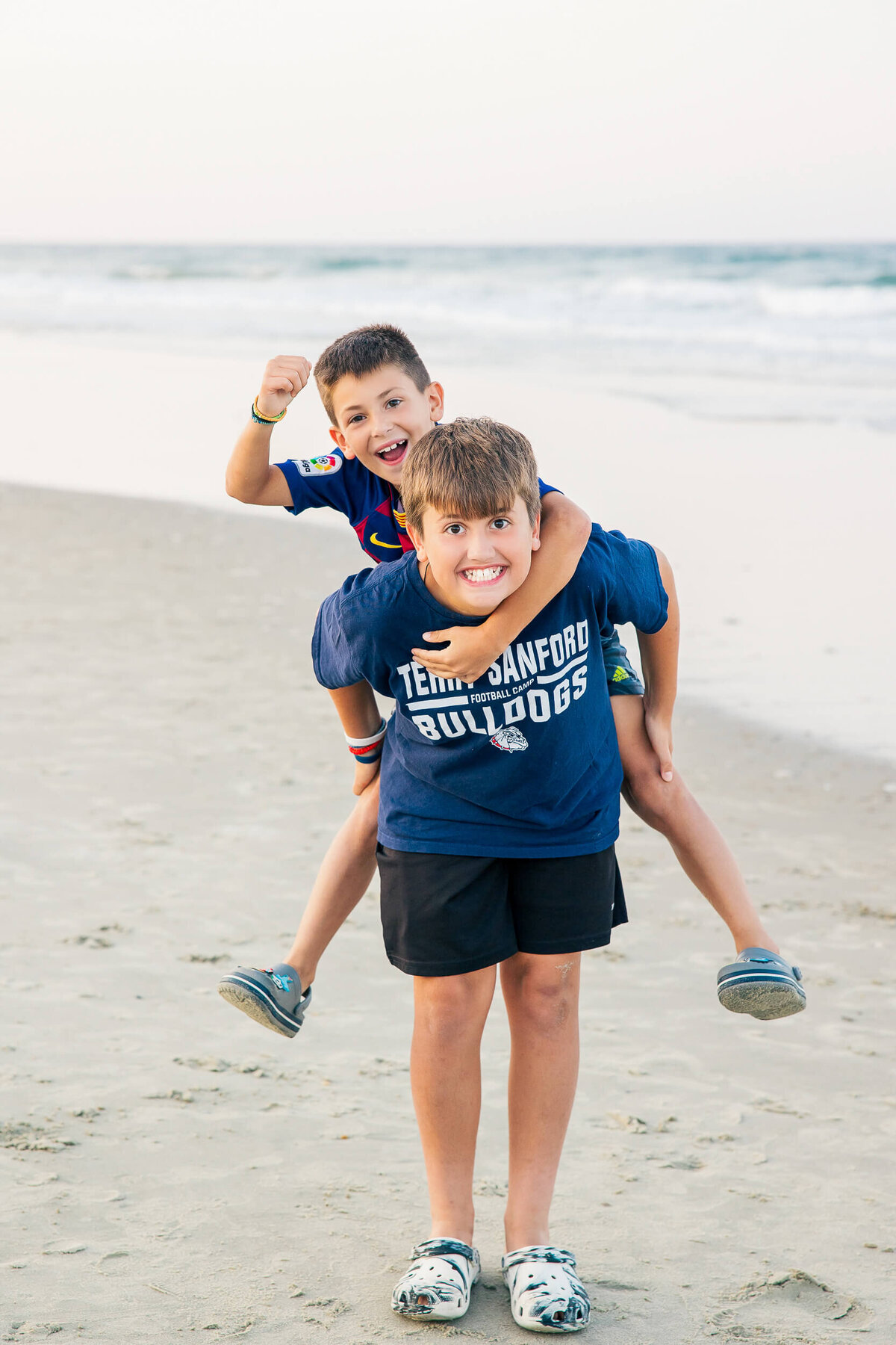 Two boys are playing on a sandy beach. The boy in front, wearing a blue "Bulldogs" shirt and black shorts, is giving a piggyback ride to the other boy in a navy shirt and shorts. Captured by a Fayetteville NC family photographer, the ocean glistens under a clear sky as their backdrop.