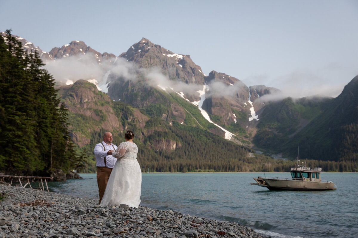 A bride and groom share a first dance on a rocky beach in Alaska as a boat sits anchored waiting for them.