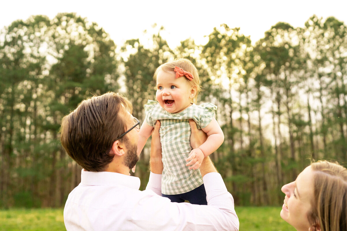 Father holding young daughter in the air while mother smiles at them.