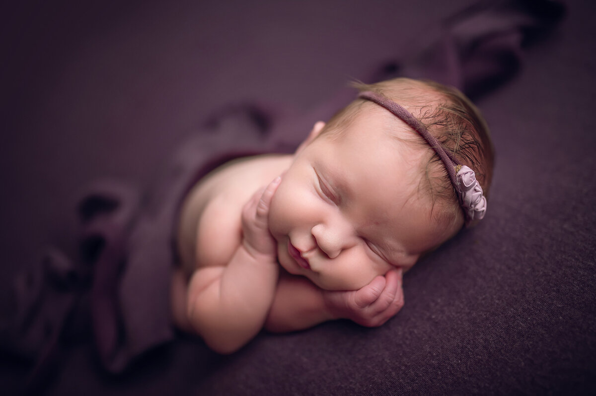 Portrait of a baby girl sleeping on a purple blanket in our Waukesha studio. She has her hands on her cheeks and is wearing a coordinating floral headband.