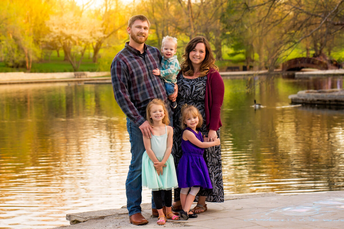 Golden hour family portrait by a lake, featuring parents and two children, captured in Gardner, Kansas. Ideal for families seeking scenic and natural outdoor photography near Kansas City.