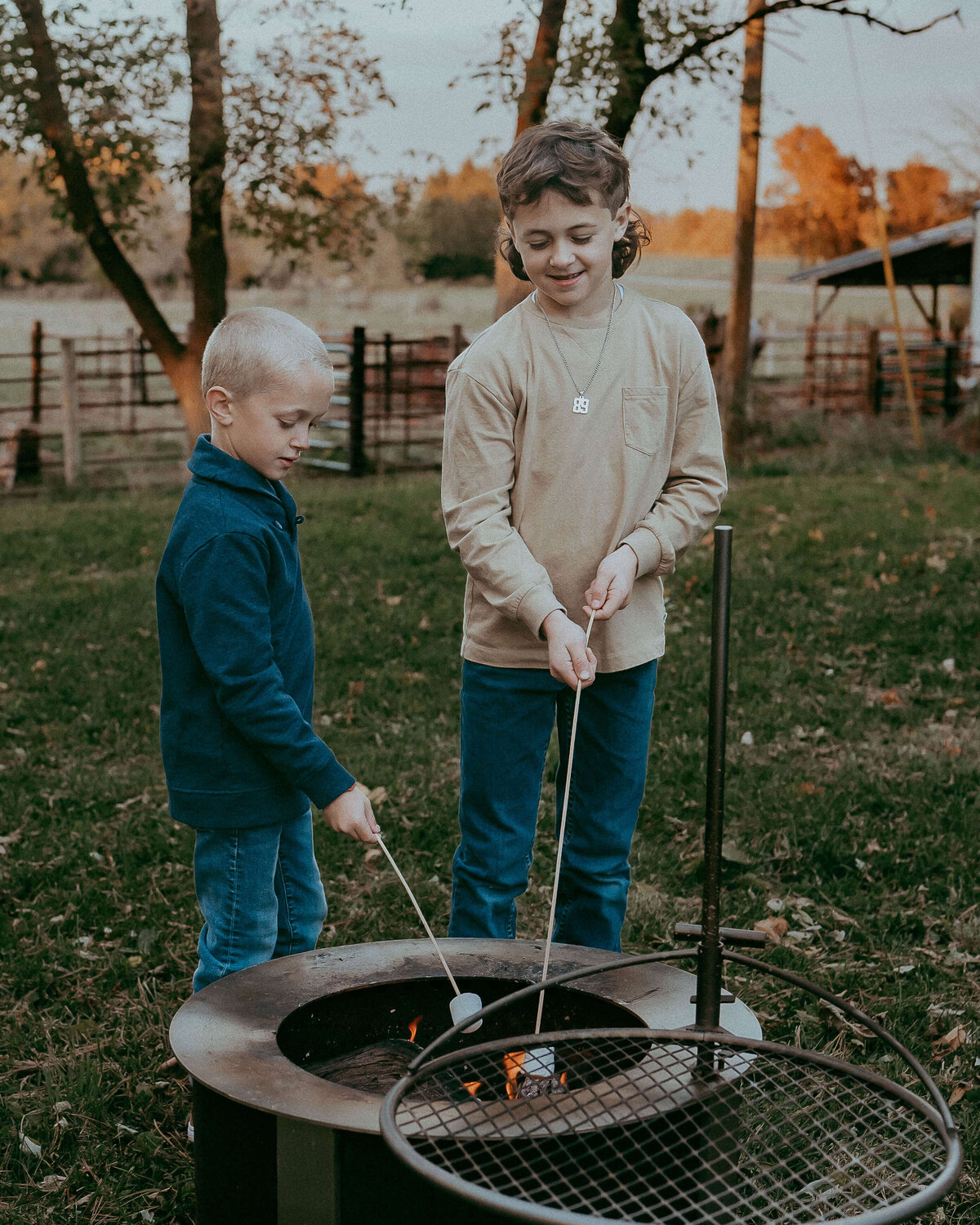 Two brothers stand at the fire pit, roasting marshmallows on their family farm as they were denim and long sleeve, neutral colored shirts.