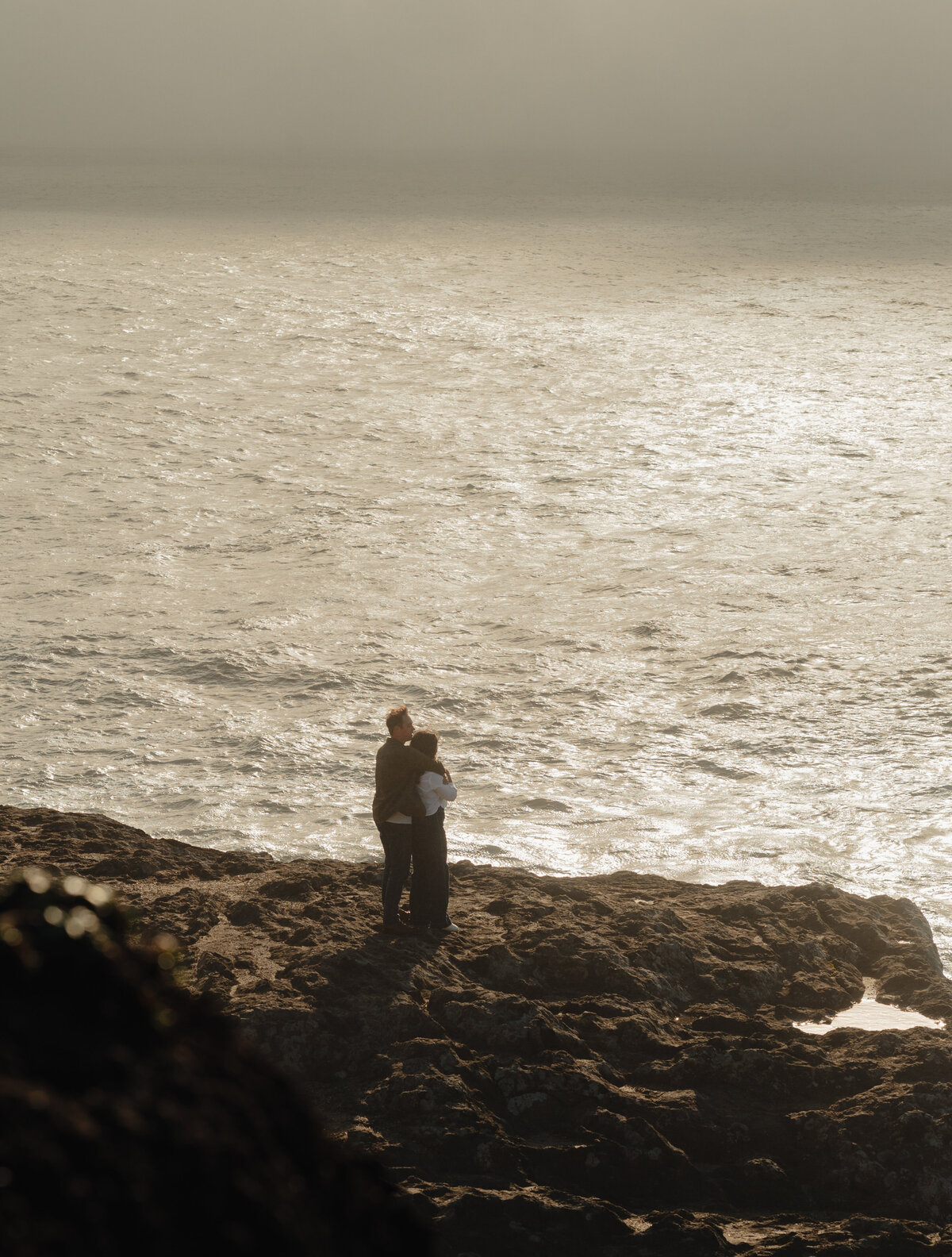engaged couple standing near the water, looking out at the horizon with golden light casting long shadows