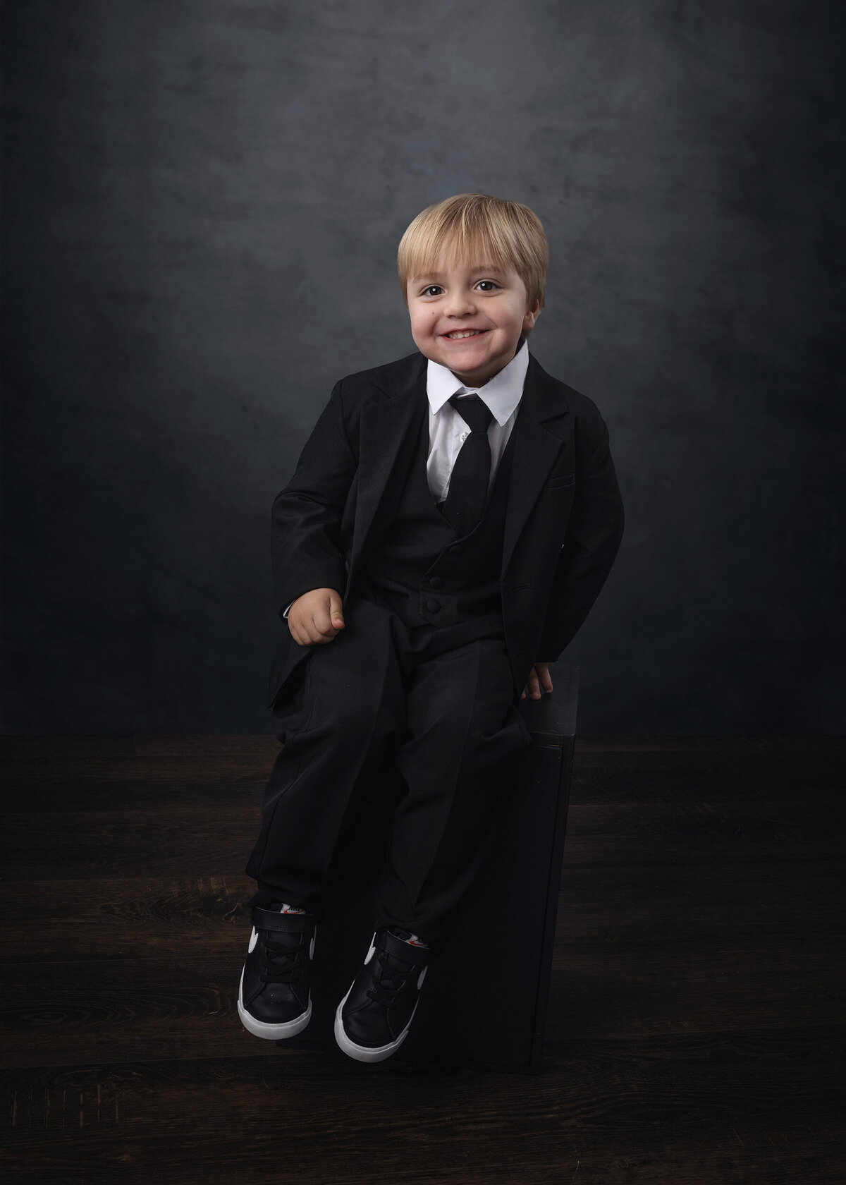A young boy sits in a Iowa Milestone Photographer’s studio in a black suit
