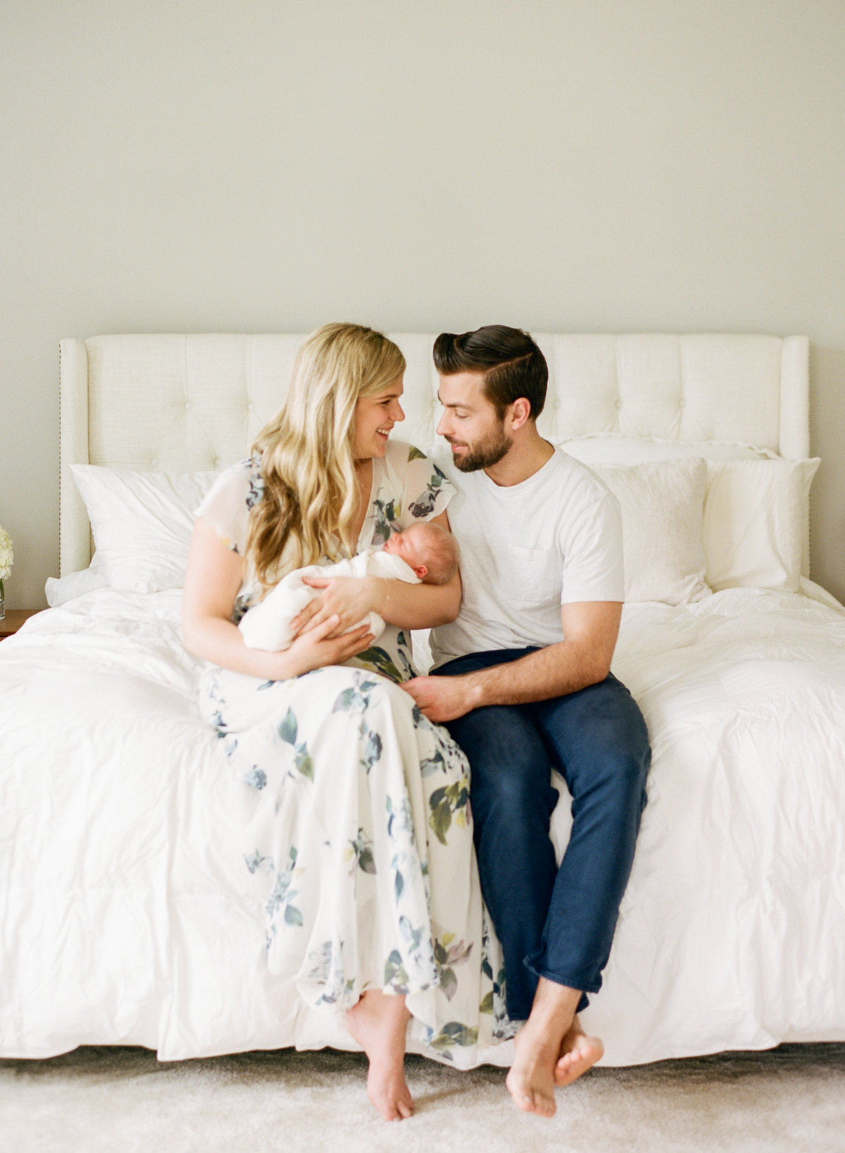 New parents smiling at their baby boy as they sit on their bed snuggling during their Raleigh newborn photography session. Photographed by Raleigh Newborn Photographers A.J. Dunlap Photography