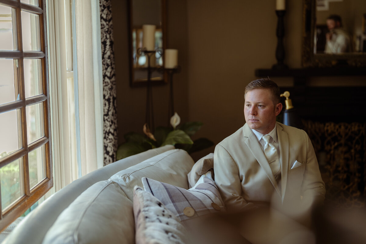 Elegant portrait of a groom sitting thoughtfully by a window on his wedding day, bathed in soft natural light. Dressed in a beige suit with a matching tie, the groom exudes sophistication in this luxury wedding portrait, captured in a beautifully decorated room.
