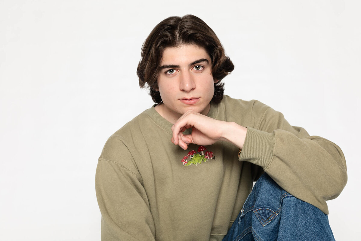 A close-up of a young male model wearing blue jeans and an olive pullover, sitting with his left knee up and his left elbow resting on his knee with his hand under his chin, looking seriously at the camera in a white studio in Tustin.