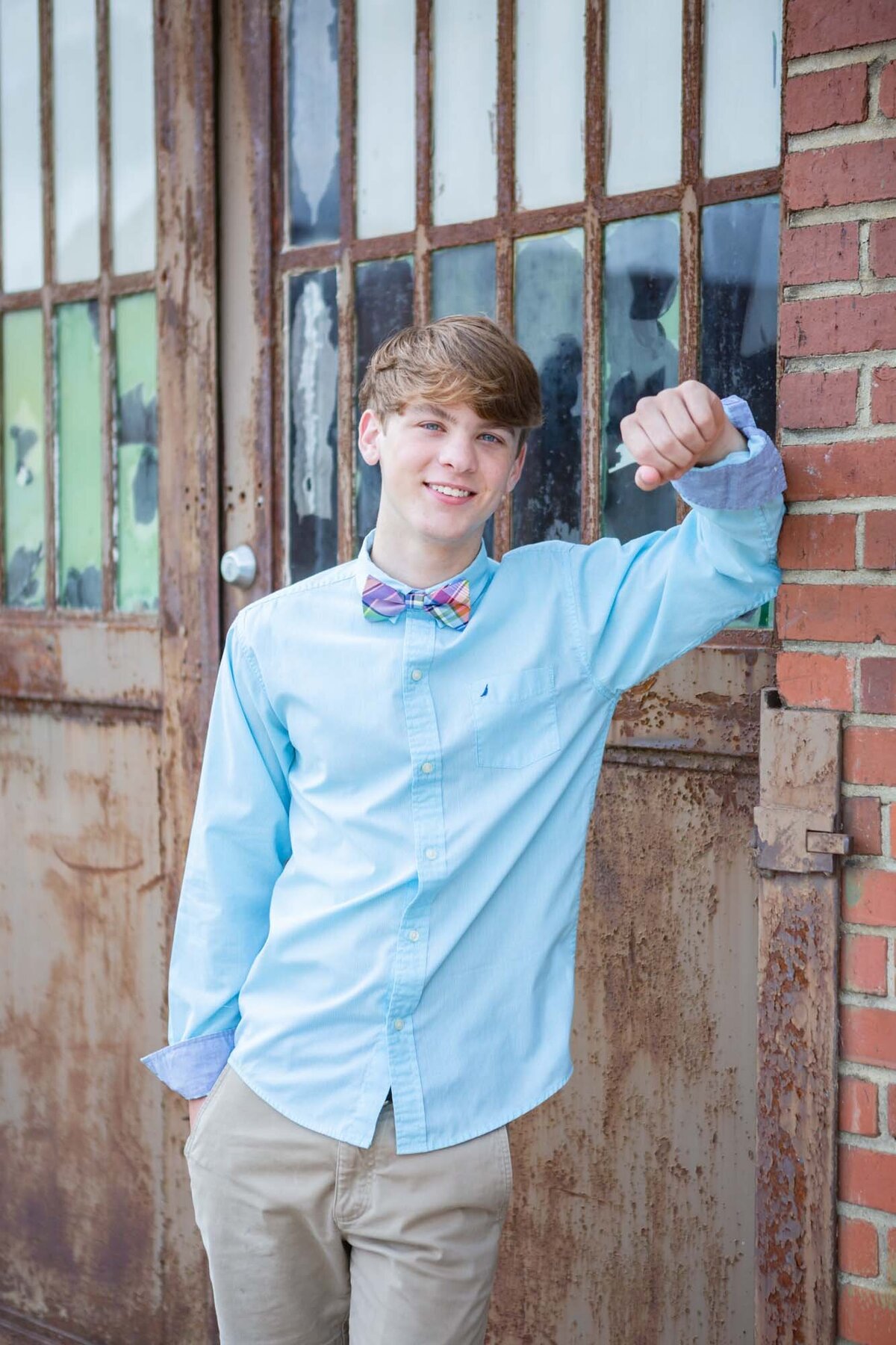Senior portrait of a guy in a blue shirt leaning against a building.