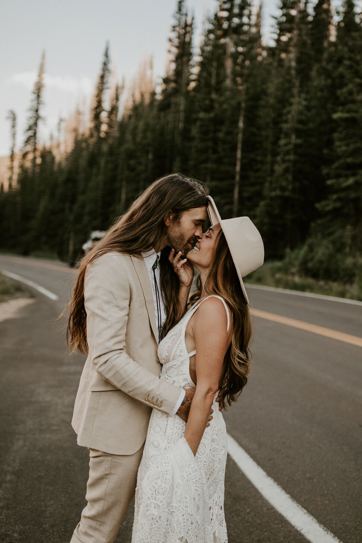Bride and groom kissing in the middle of a road in the mountains, wearing an ivory suit and white wedding gown with an ivory hat.