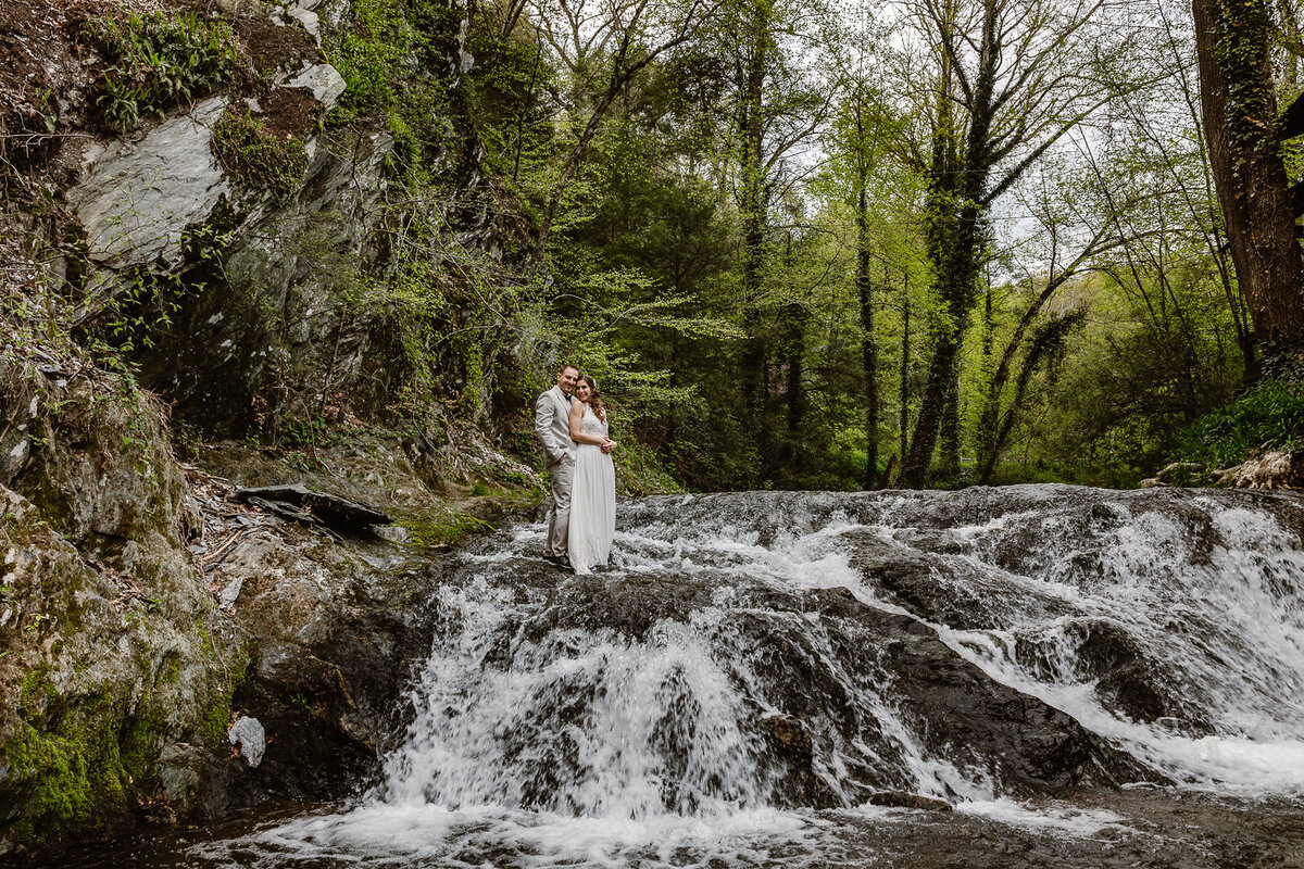 Matt holds Danielle from behind while they stand on a waterfall.