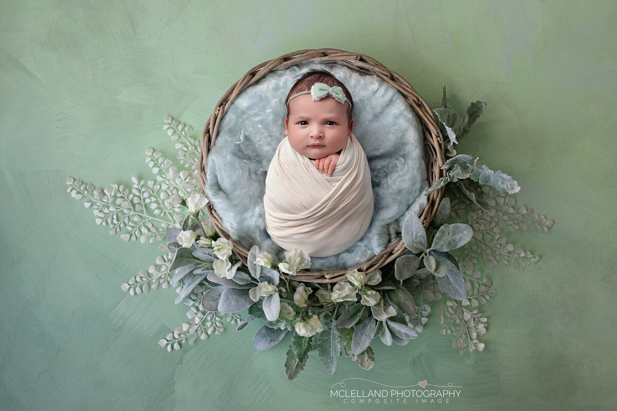 Newborn baby wrapped in white posed in a botanical wreath with blue-green florals.