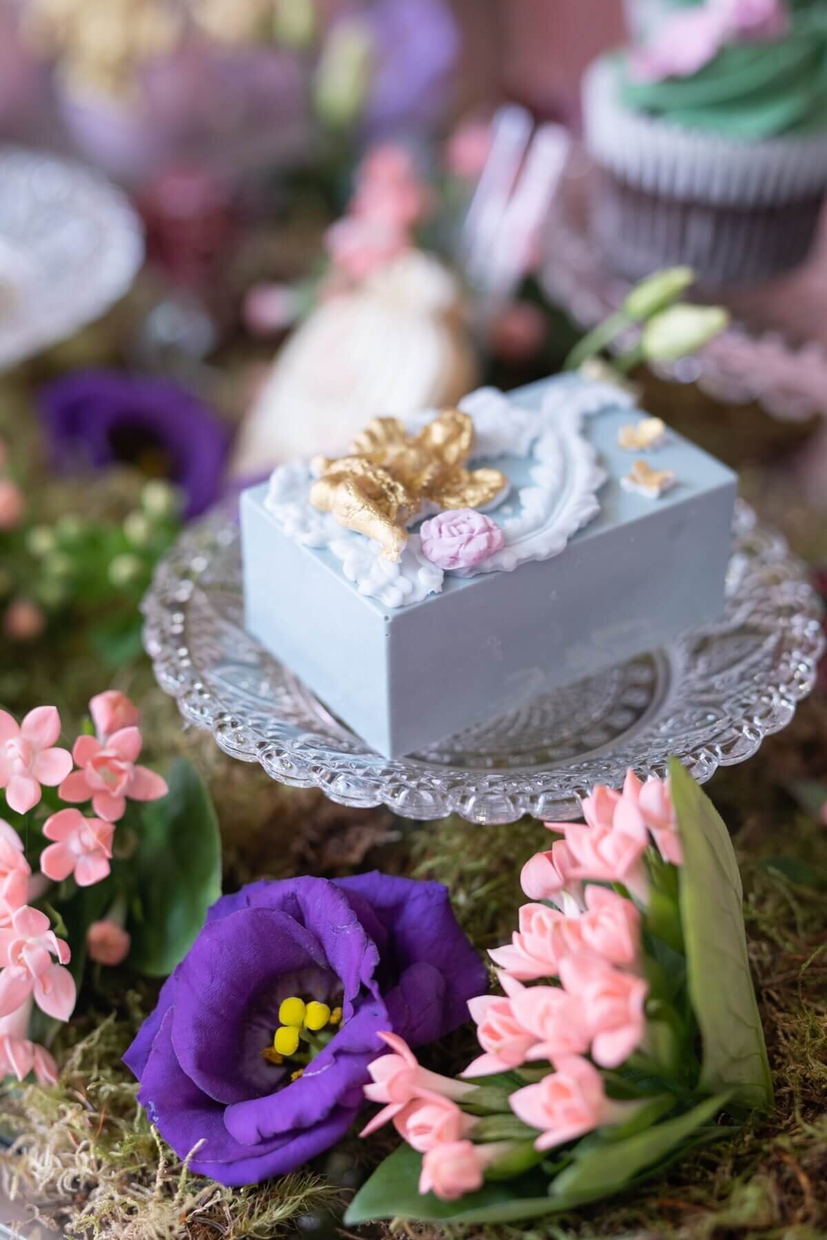 Small blue cake with gold sugar art on small glass plate with purple and pink flowers