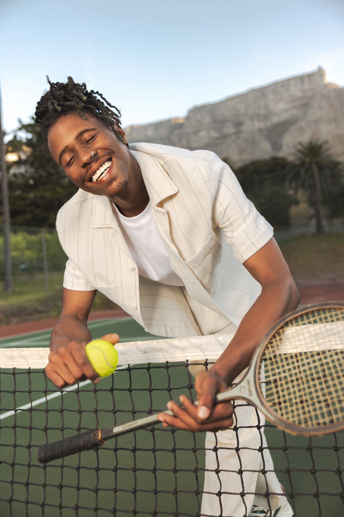 Smiling man leaning over a tennis net while holding a tennis racket and ball, wearing a cream striped outfit with Table Mountain in the background.