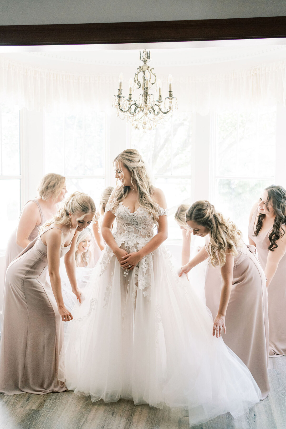 A bride in a white wedding dress stands in the center while bridesmaids in pink dresses arrange her gown in a brightly lit room with a large window and a chandelier overhead, capturing the essence of a modern &amp; romantic wedding in Calgary at Norland Historic Estate.