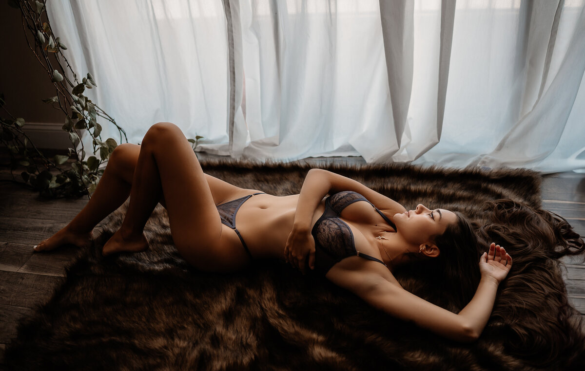 Elegant boudoir photography featuring a dark-haired woman in lace lingerie posing gracefully on a faux bear skin rug, with soft natural light streaming through sheer curtains in Tampa, Florida.