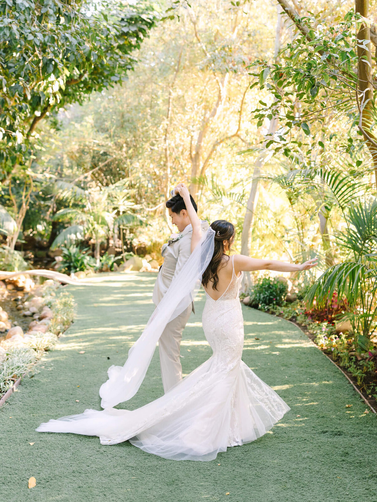 A couple dances on a sunlit garden path, surrounded by lush greenery. The bride's elegant veil and gown cascade gracefully.