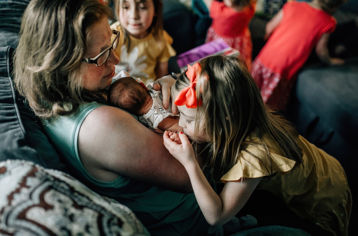 Mom holding newborn baby with young girl kissing the newborns hand.