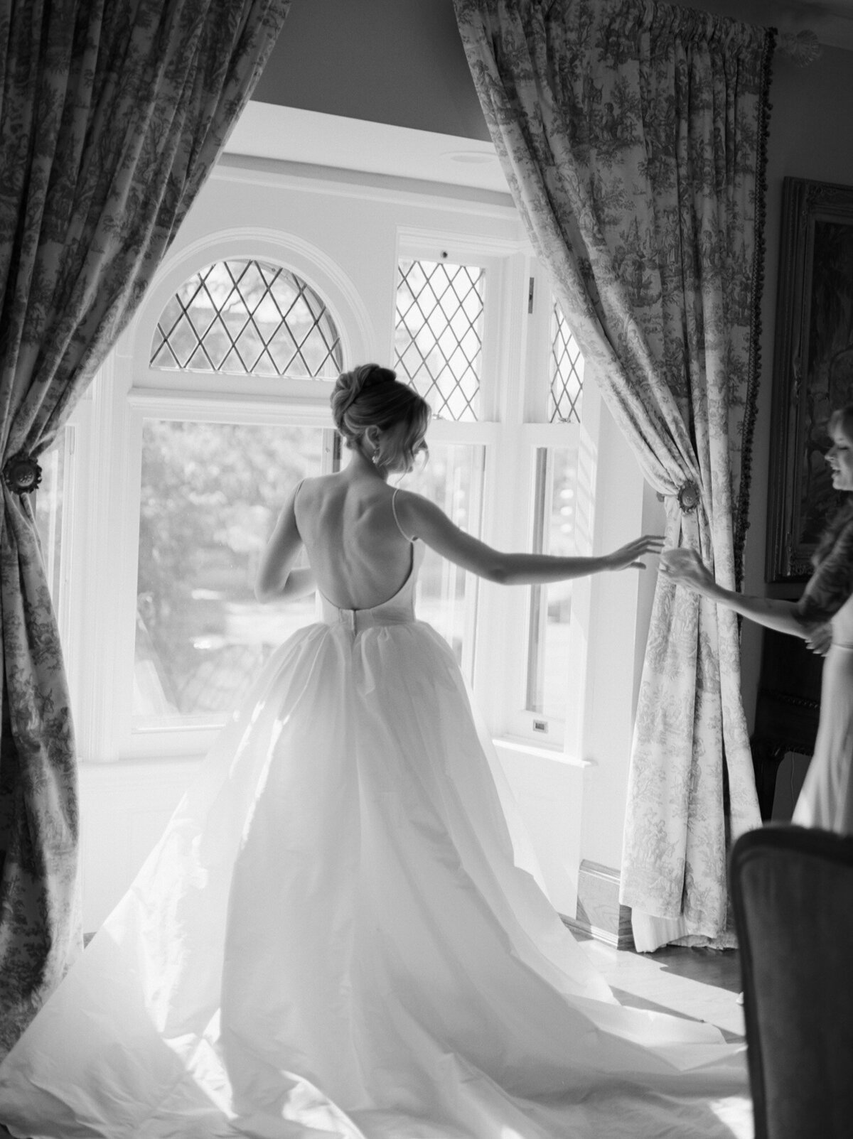 a bride in open-back ballgown in morning window light reaches for perfume bottle