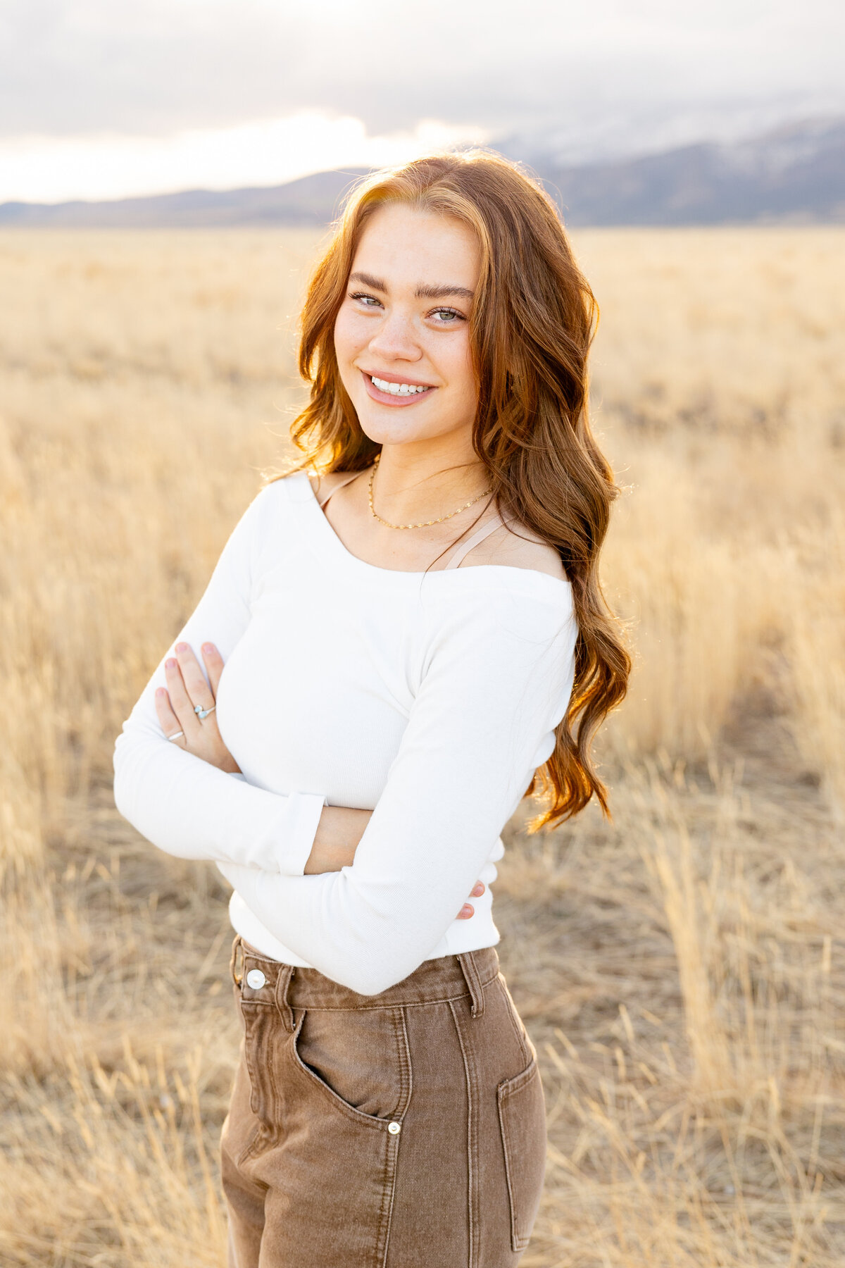 A young woman with long auburn hair smiles confidently in a white long-sleeve top and brown pants, standing in a sunlit field in Eagle Mountain, Utah, with distant mountains behind her.