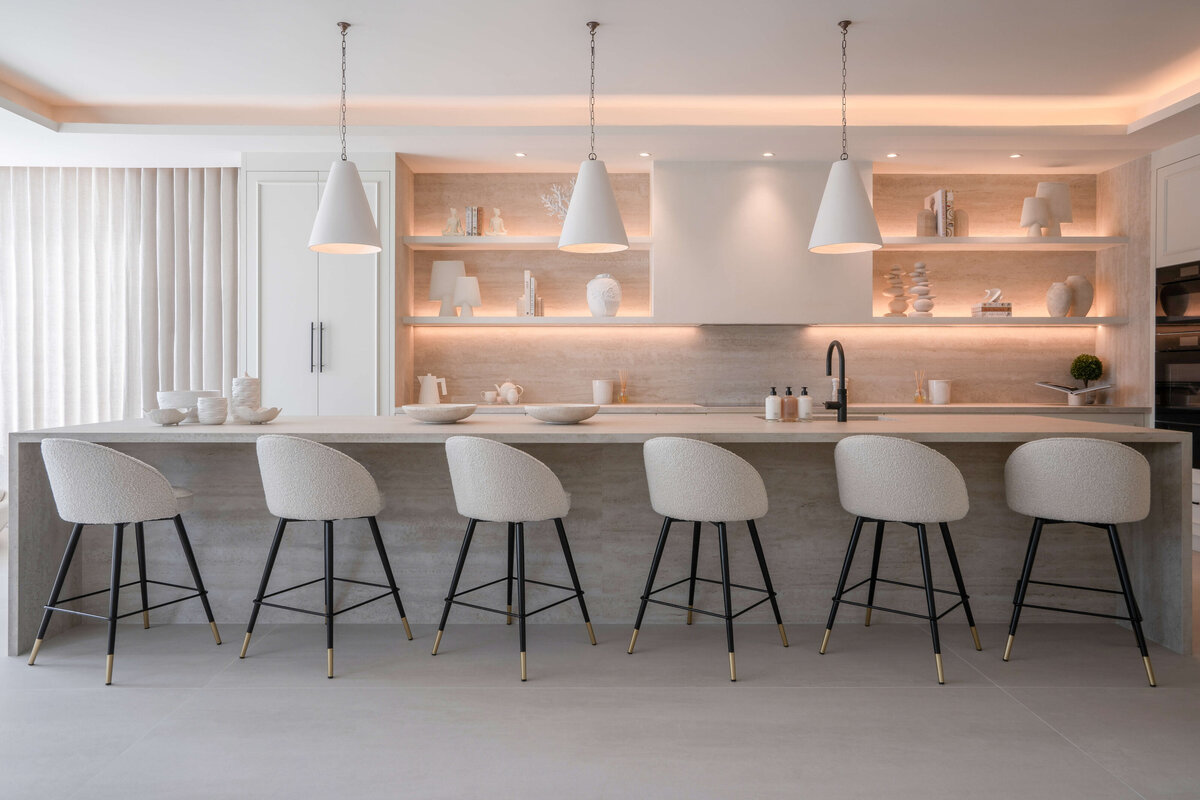 A large, open kitchen decorated with a bright, minimal aesthetic. Along a long bar sit six white rounded barstools with black legs. Above are three white pendant lights. Behind, there  is built-in white shelving illuminated by soft integrated lighting and decorated with minimal white chic decor.