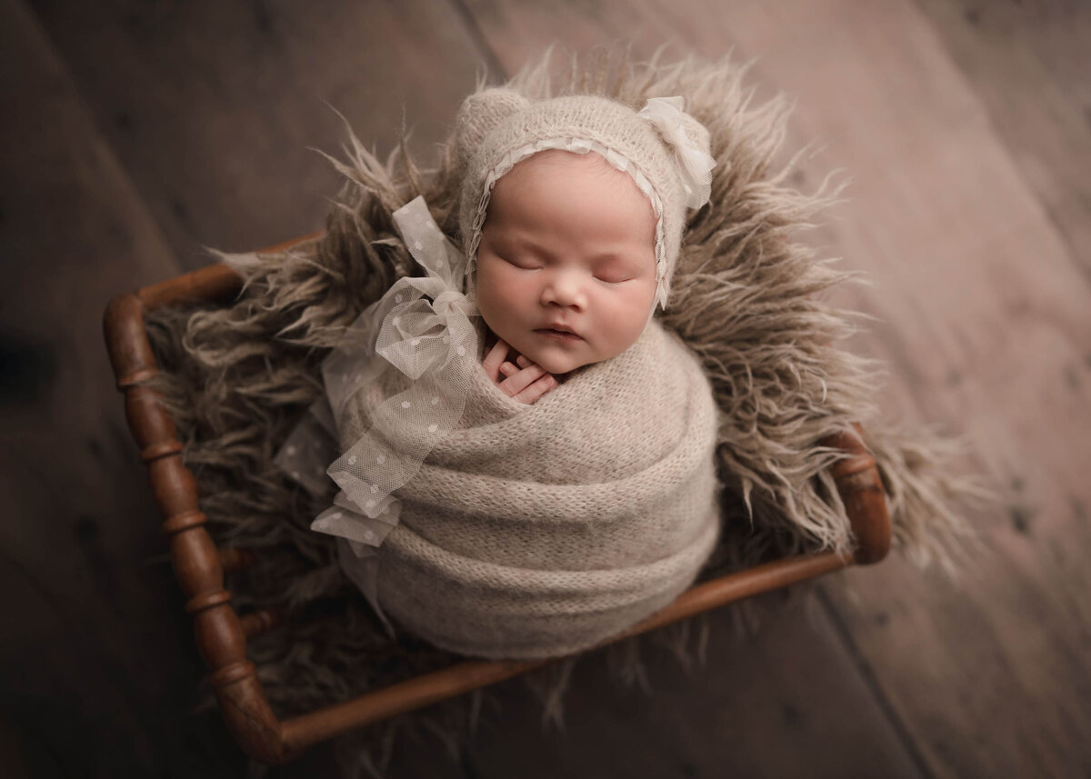 Aerial image. Menifee newborn photoshoot. Baby girl is wrapped in a light beige swaddle with her fingers peeking out. She is wearing a matching bonnet with small bear ears and a trailing ribbon to fasten it. Captured by best Menifee newborn photographer Bonny Lynn Photography
