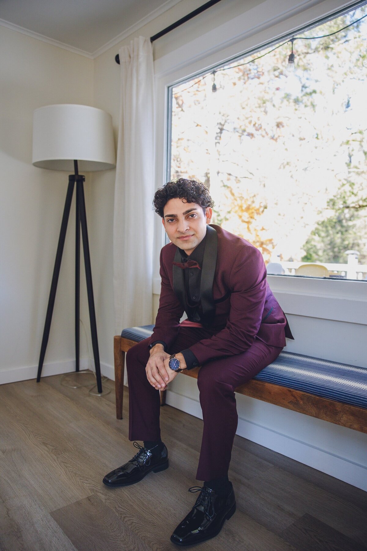 Groom sitting indoors before the ceremony, dressed in a maroon suit and black shirt.