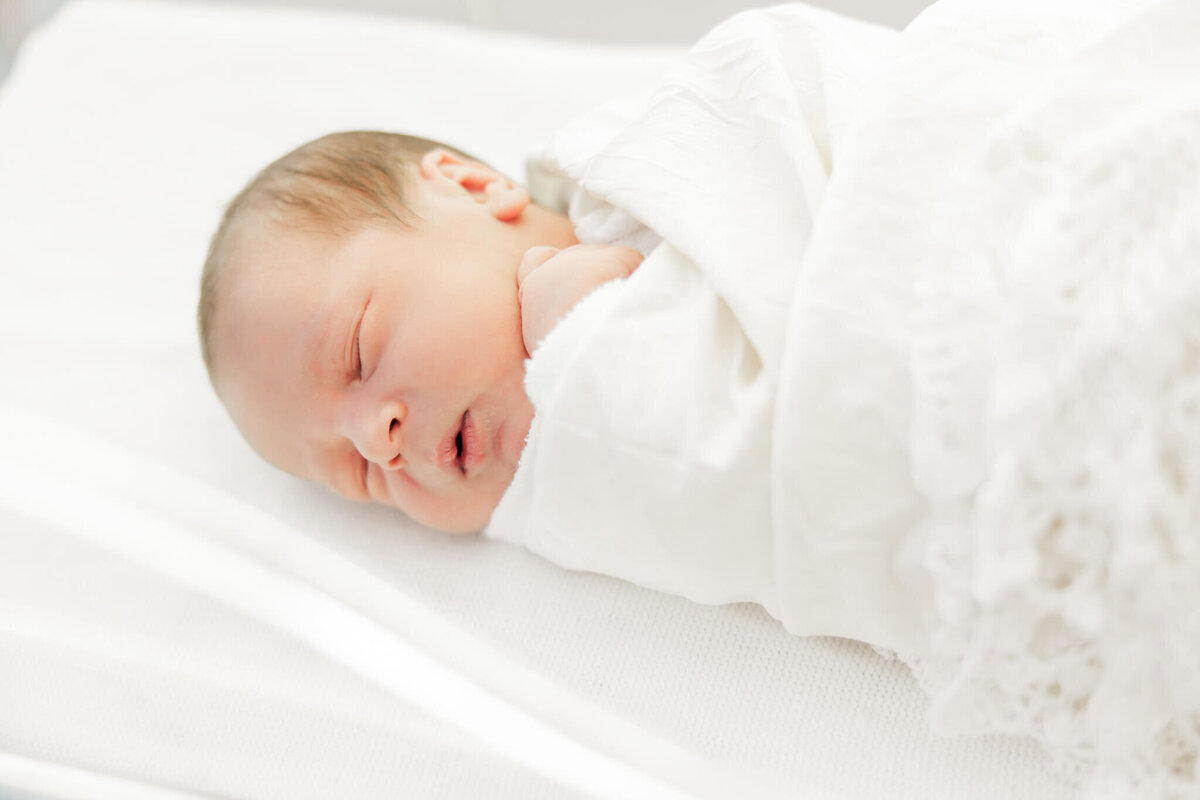 Newborn baby swaddled in white, lace blanket, asleep in a hospital crib for a Fresh 48 photo session. Captured by Julie-Anne Photography.