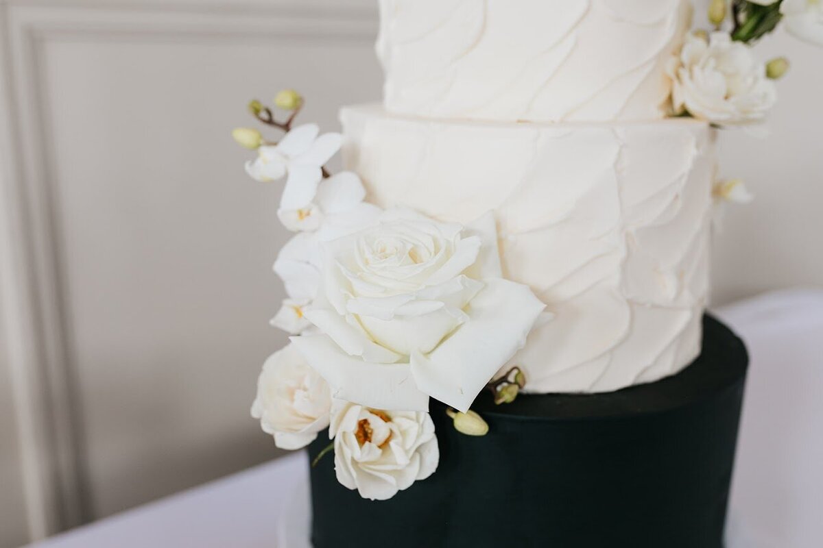 A close up of a three tier cake. The bottom tier is smooth black frosting, and the top two tiers are textured white frosting. Decorated with clusters of fresh white flowers.