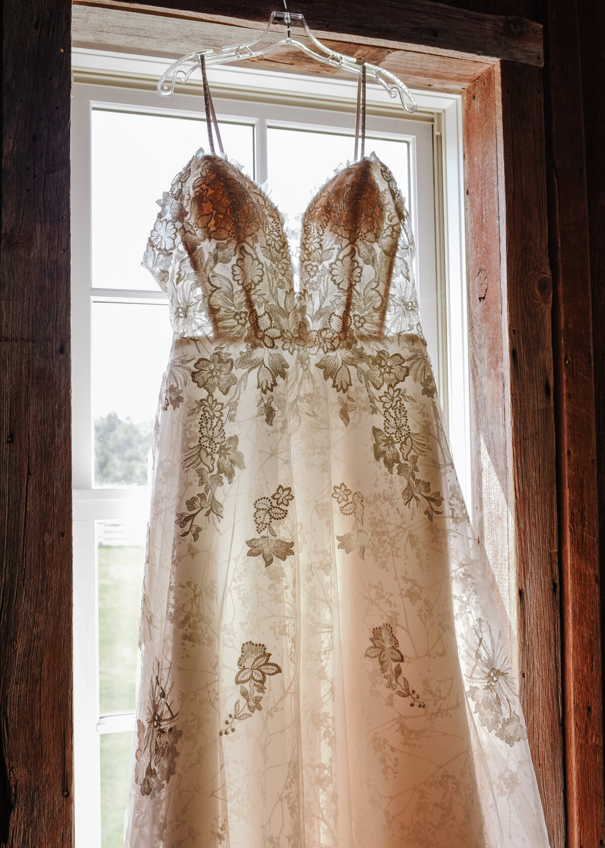 An elegant wedding dress adorned with lace details hangs in front of a window, bathed in soft natural light, awaiting the special moment it's worn by jen Jarmuzek photography a Minneapolis wedding photographer