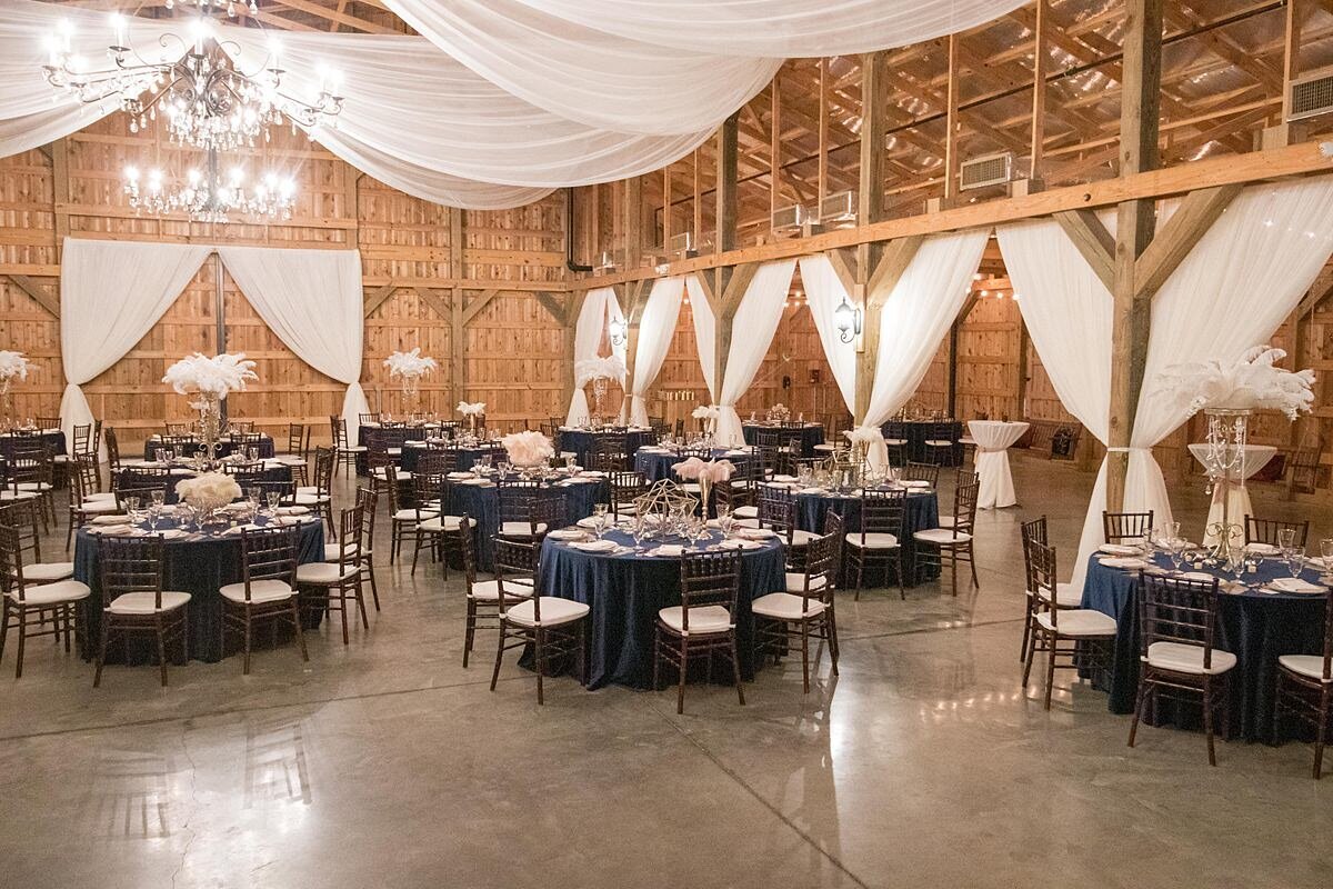 Navy velvet table cloths and brown chiavari chairs with white cushions are set throughout the reception barn at Saddle Woods Farm. The tables are set with crystal and gold centerpieces and ostrich feathers. The walls and ceiling have been decorated with sheer ivory drapery and are accented by large gold and crystal candelabra chandeliers.