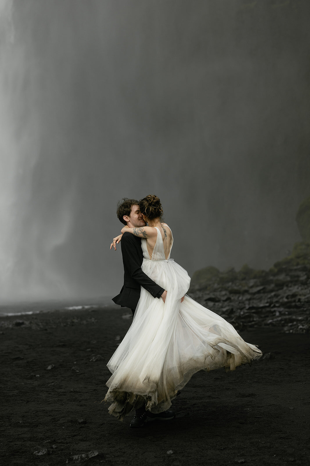 Groom holding bride as her wedding dress swirls in front of a dramatic Iceland waterfall, captured by a destination elopement photographer.