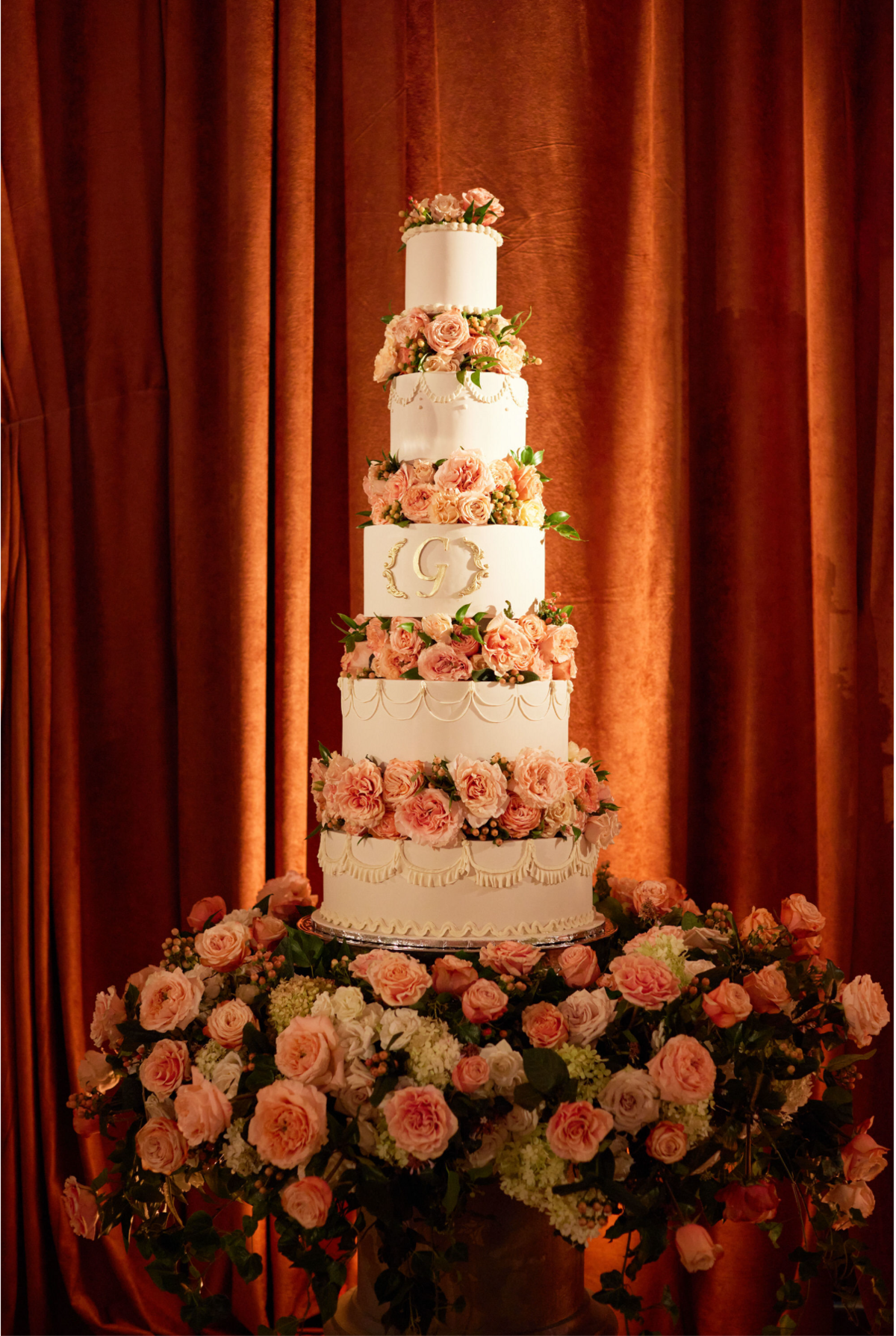 This stunning photo was taken at the renowned Willard Intercontinental in Washington D.C., known as one of the top wedding venues and hotels in the city. The talented photographer Shawn Connell captured the elegance of the scene perfectly. The focal point of the image is a tall, elegant five-tiered white wedding cake, exquisitely adorned with intricate floral decorations and a monogram at the center. The cake is displayed on a table surrounded by a lavish arrangement of pink and white flowers, creating a romantic and luxurious ambiance. The backdrop of rich, reddish-brown curtains adds a touch of sophistication to the overall setting. Clayton Films can be added as the Videographers for this event.