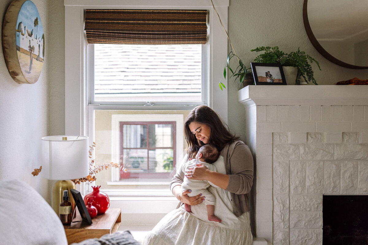 mom in off-white dress  holding newborn in off-white outfit sitting in window seat