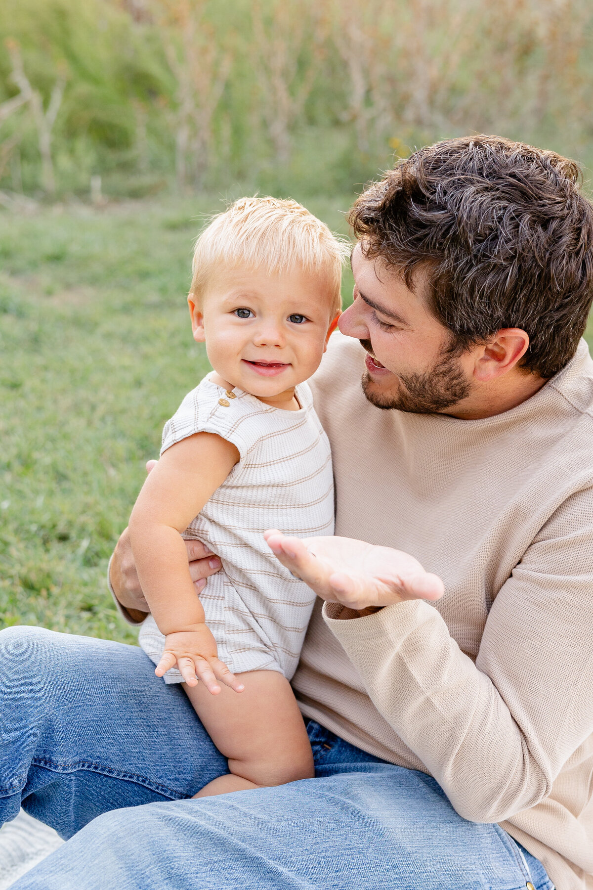 A loving father in a beige sweater sits on the grass, holding his blonde baby boy in his lap, sharing a joyful moment in Eagle Mountain, Utah. Captured by Cassie Lee Photography.