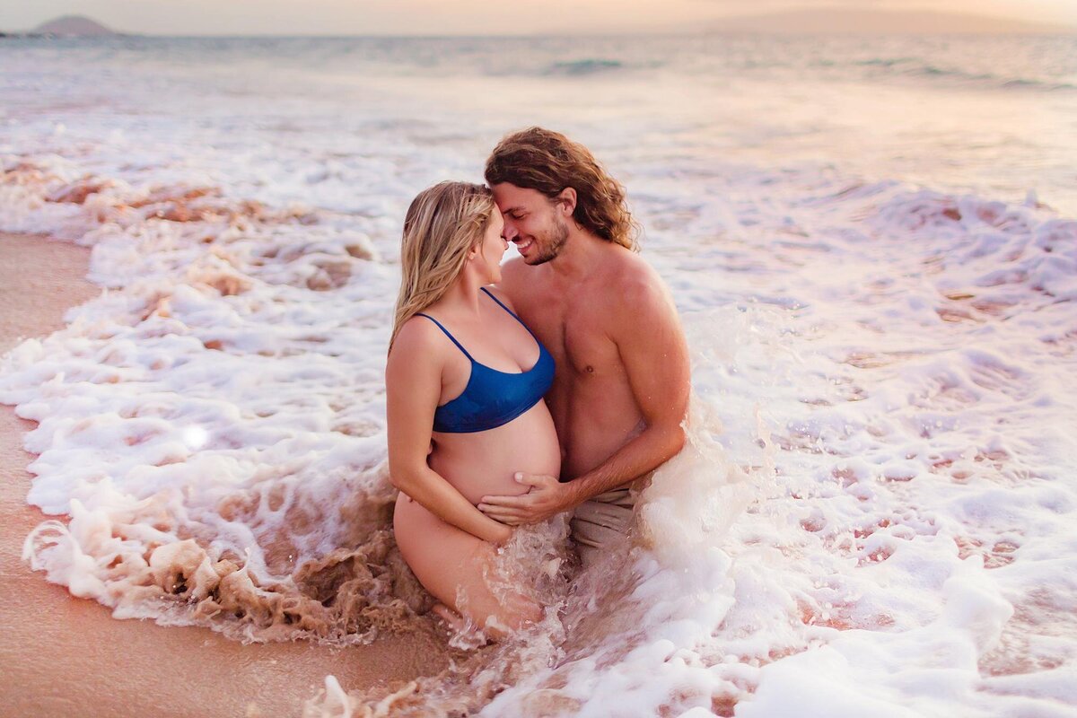 Couple kneels and embraces on the sand as a wave washes over them during a romantic babymoon portrait session in Maui.