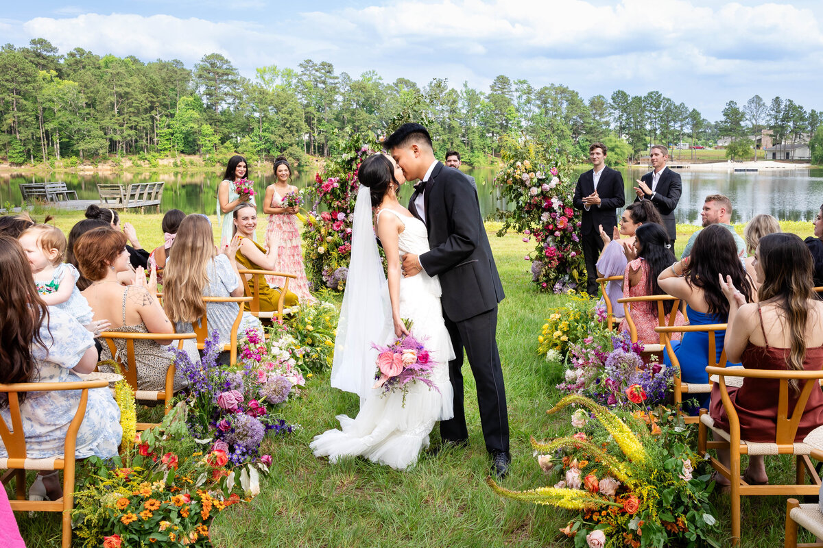 Experience the magic of an outdoor wedding by the lake, featuring a joyful couple surrounded by vibrant floral arrangements and smiling guests. This picturesque setting offers the perfect inspiration for a romantic and colorful celebration.