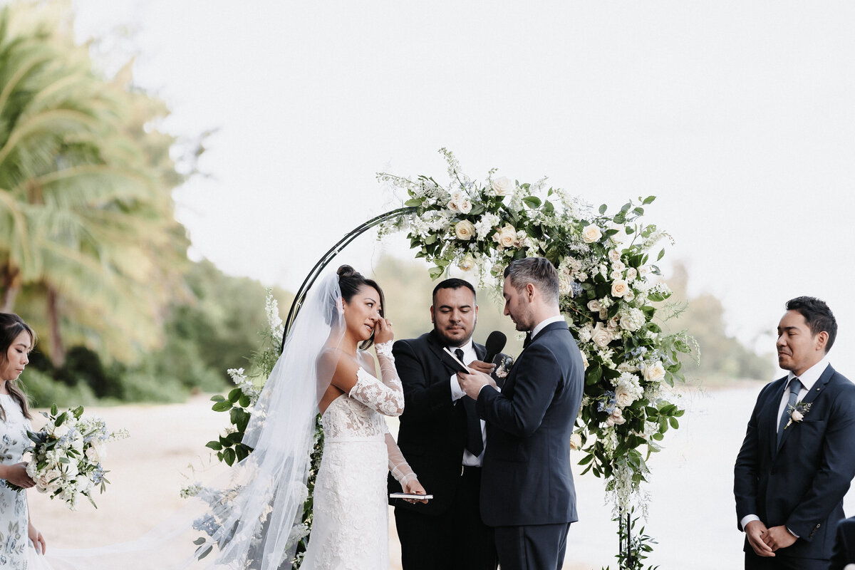 Ceremony on the beach with floral arch behind couple, bride is wiping away a tear as the groom holds the microphone reading vows