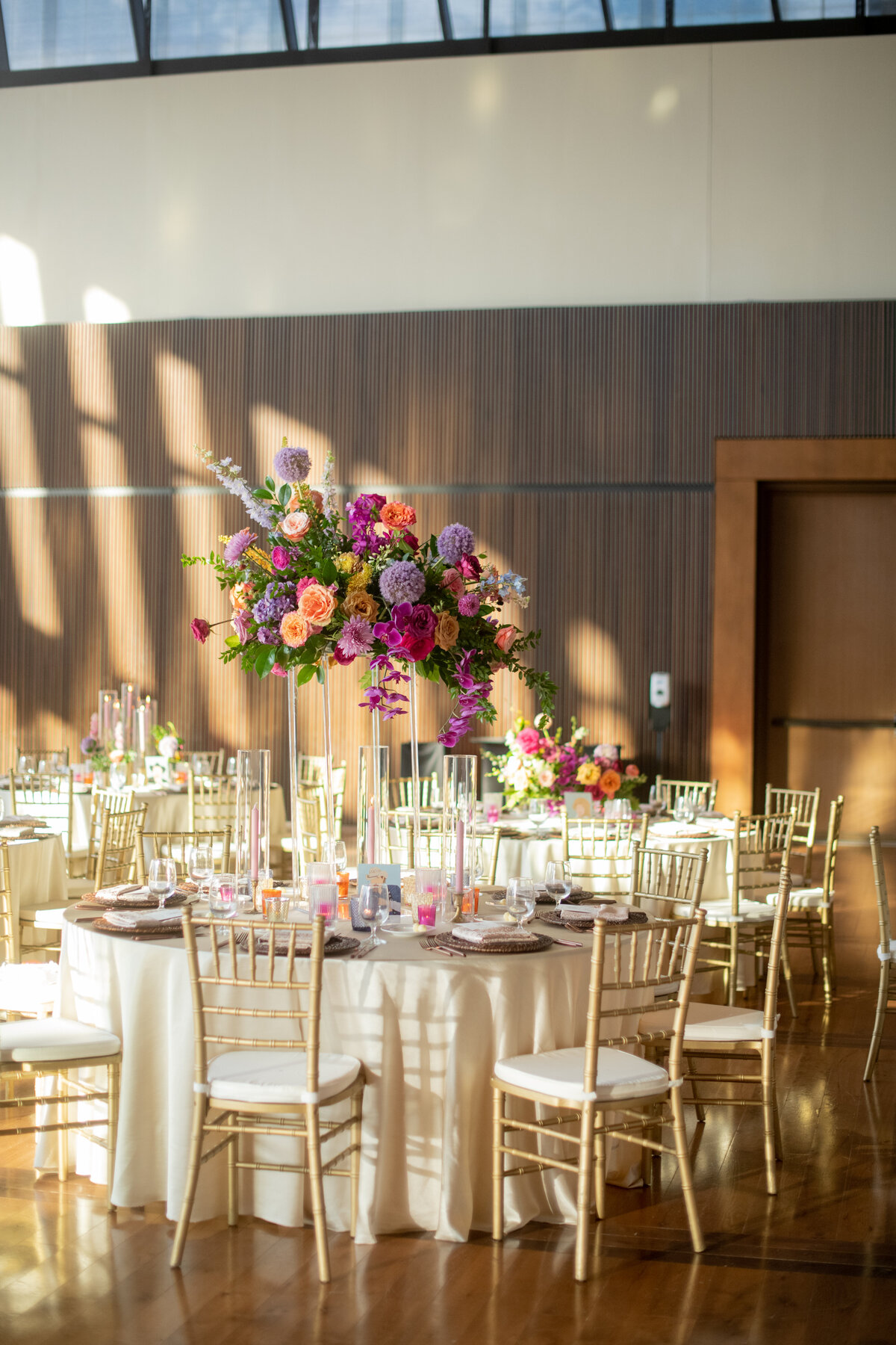 Lush elevated centerpieces in sunset hues of rosy pink, lavender, orange, and golden-yellow composed of phalaenopsis orchids, petal heavy roses, allium, delphinium, eremurus and natural greenery. Design by Rosemary and Finch in Nashville, TN.
