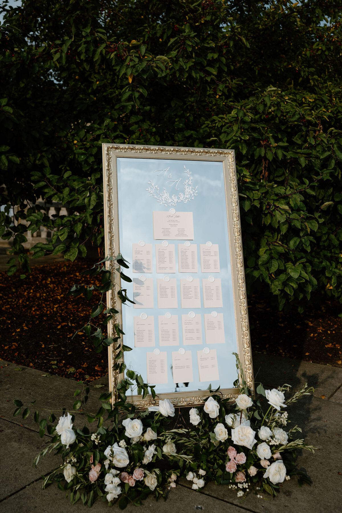 Elegant wedding day stationery display with gold-framed mirror seating chart, floral crest, and guest list cards.