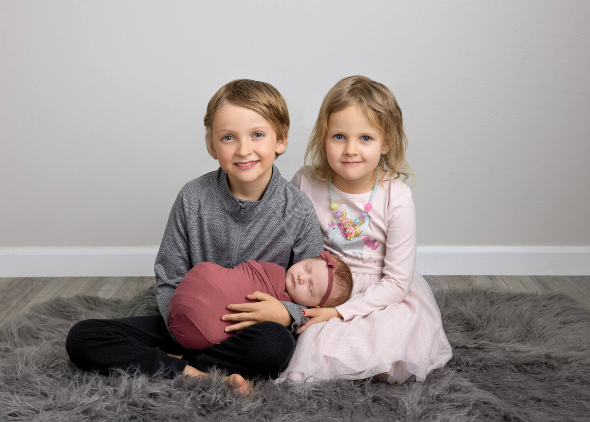 brother and sister sitting holding their newborn baby sister on a grey shaggy rug