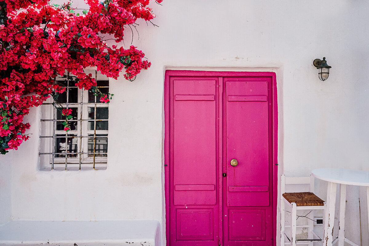 White walls with a bright pink door and pink Bougainvillea flowers climbing up the wall
