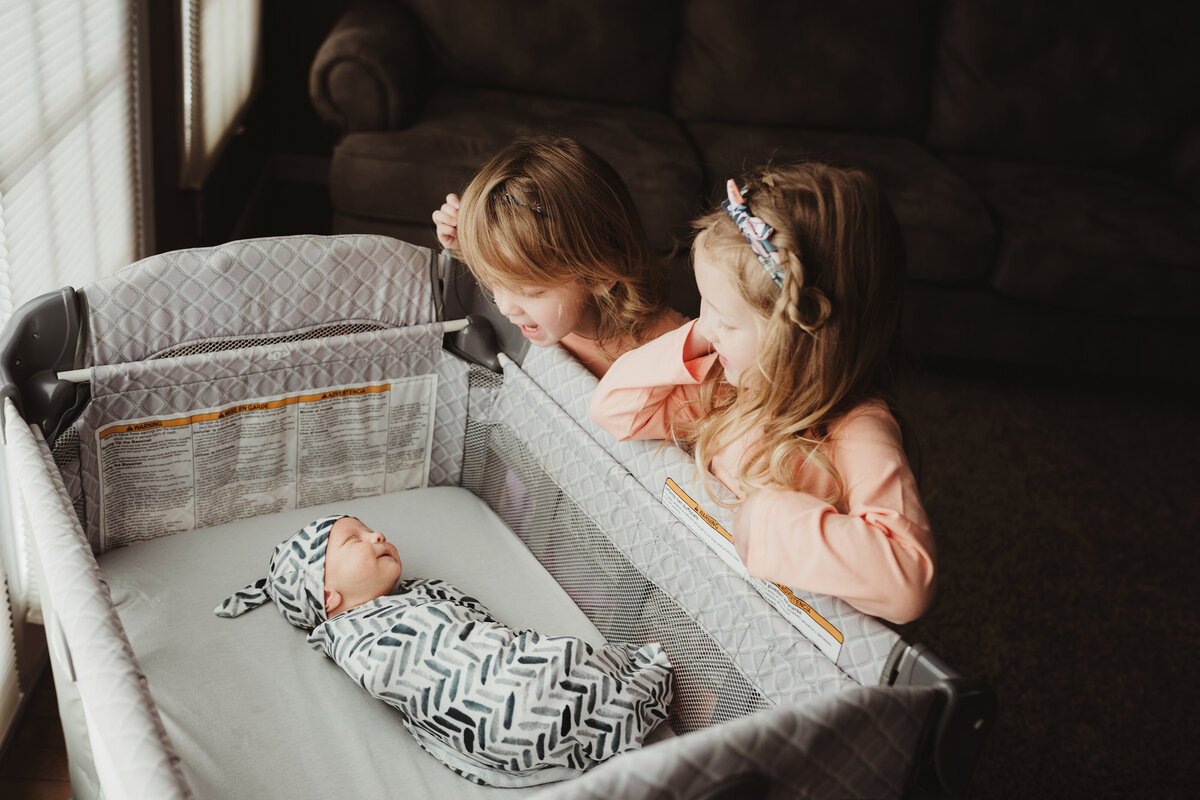 Two big sisters smile back at their baby brother lying in the bassinet.