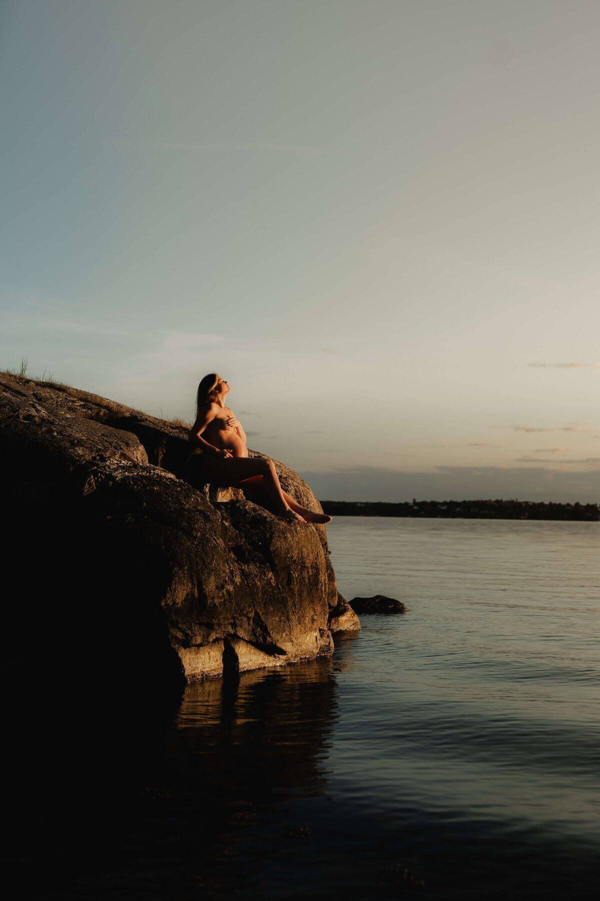 Bild på en gravid kvinna sittandes långt ifrån på ett berg vid vattnet under en gravidfotografering i Stockholm