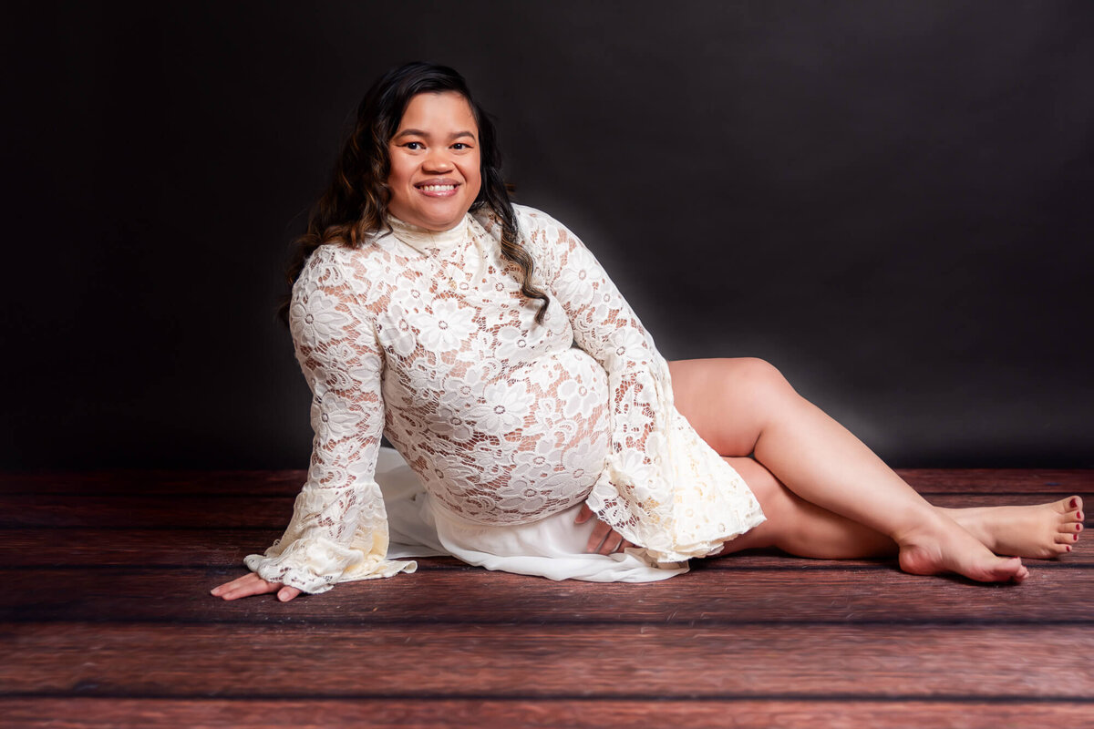 A pregnant woman in a flowing white dress sits gracefully on a wooden floor, radiating calm and beauty.