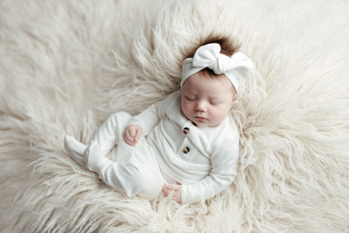 Studio newborn photography - sleeping newborn baby girl with lots of brown hair. She is wearing a white onesie and a matching tied white headband on a white flokati.
