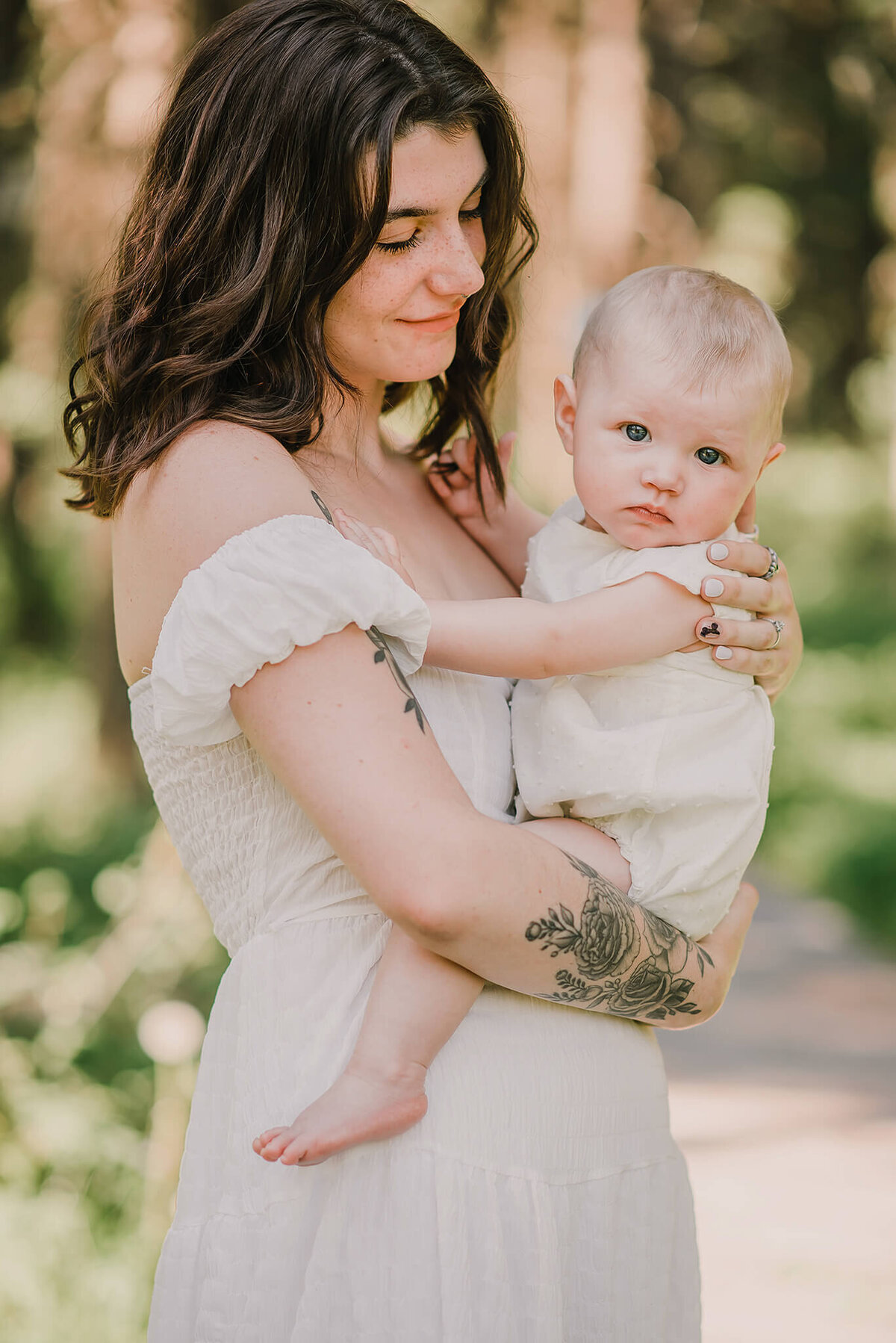 An up close portrait of a beautiful mother holding her infant daughter on her hip. Baby girl looks at the camera while mom smiles down at her. They are in alush green forest. Taken by Brittany Danielle
