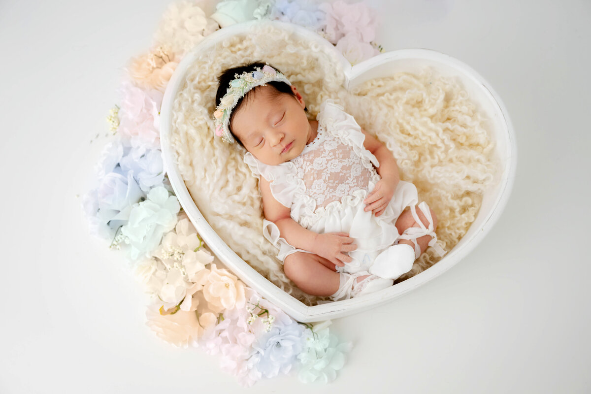 Newborn baby in an all white dress and little ballerina shoes, sleeping in a heart shaped prop with rainbow flowers on the outside.