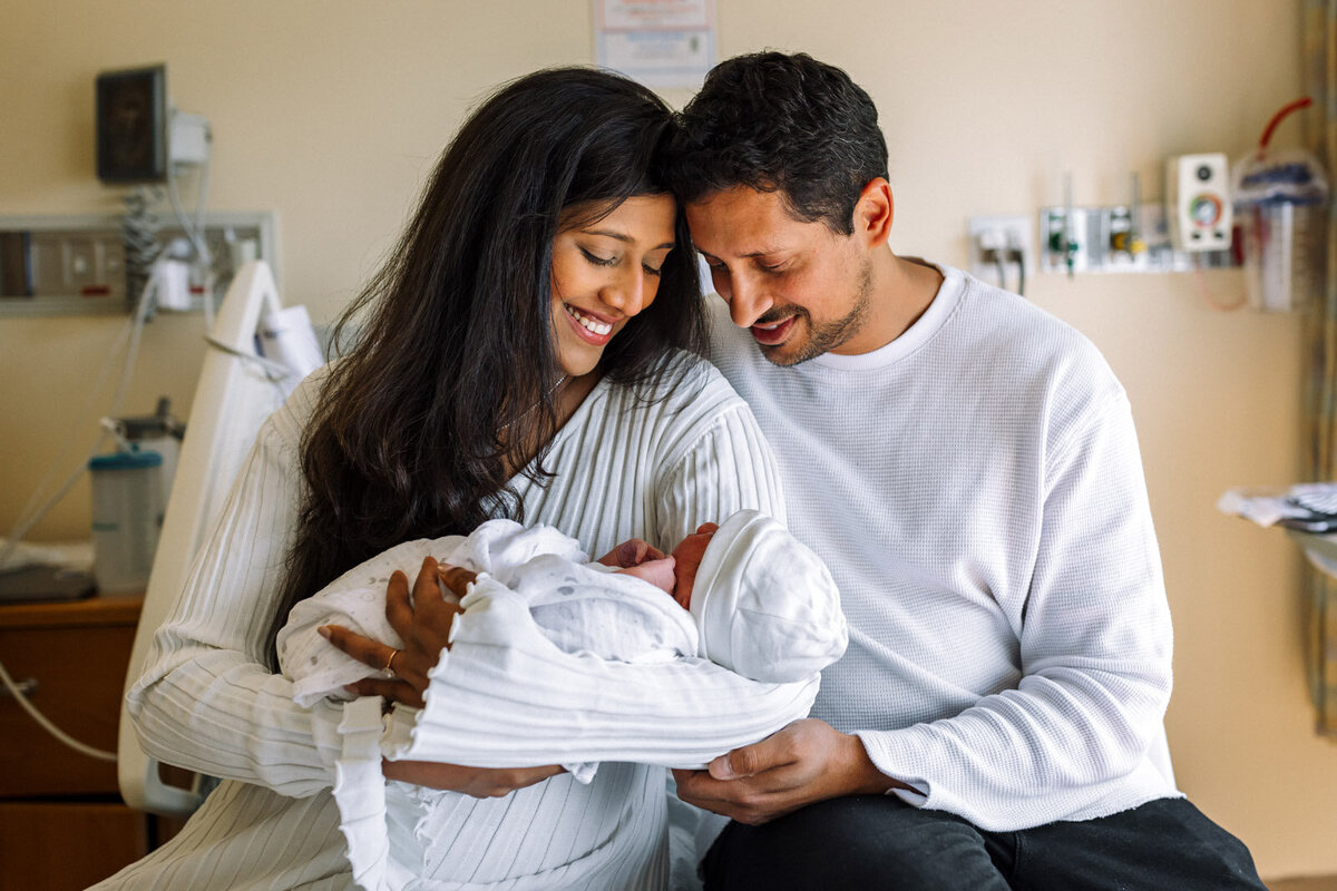 parents sitting in hospital bed holding swaddled newborn and looking down at him smiling