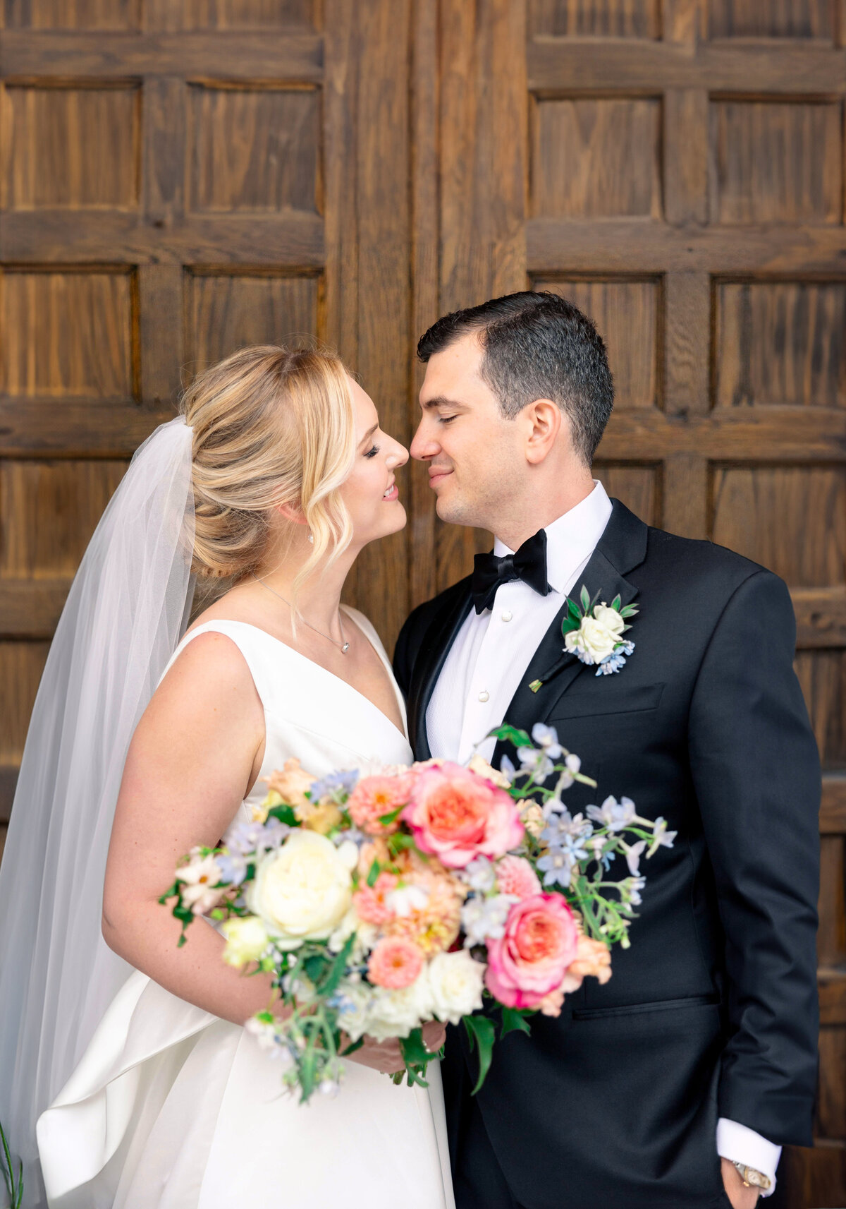 A bride and groom stand closely in front of wooden double doors. The bride holds a colorful bouquet and wears a white gown and veil, while the groom is in a black tuxedo with a boutonniere. They gaze into each others eyes lovingly.